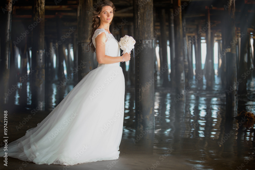 Gorgeous artistic portrait of beautiful bride in white dress holding bridal bouquet under pier