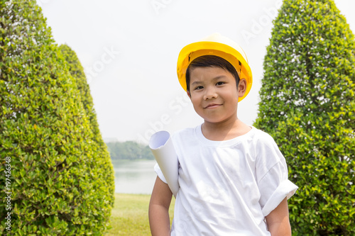 Asian boy in white t-shirt with yellow helmet dress like engineer and hold roll of paper with garden scene