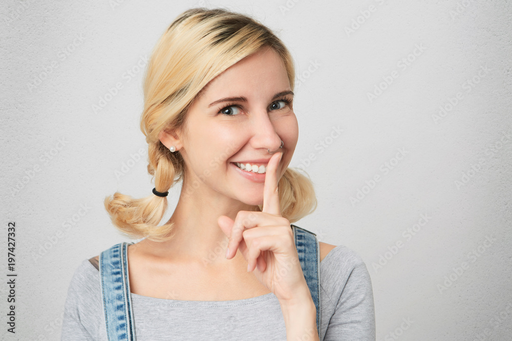 Portrait of funny excited blonde female keeping finger at lips, having mysterious look, asking not to give away her secret. Isolated over white wall. Portrait of funny excited blonde female keeping finger at lips, having mysterious look, asking not to give away her secret. Isolated over white wall.