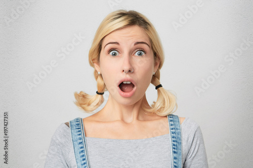 Isolated shot of young attractive female stares at camera with shoked expression, recieves news, keeps lips rounded.