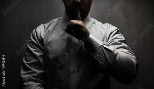 Photography Photo of a scary horror man in white shirt standing under upper light on black background showing silence hand sign