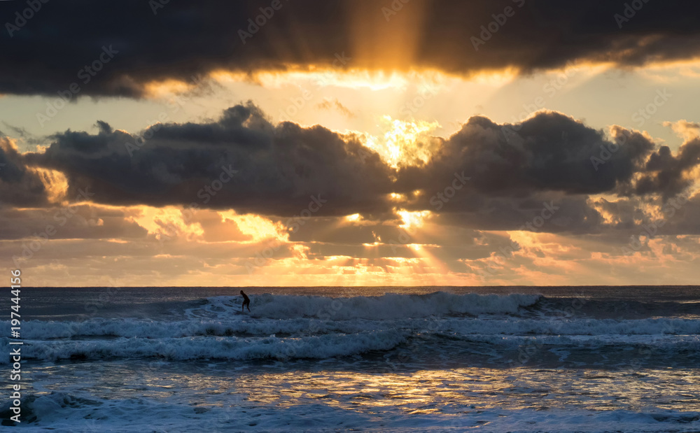 Dramatic sky with sunburst rays through clouds as single surfer rides waves in the early morning on secluded beach.