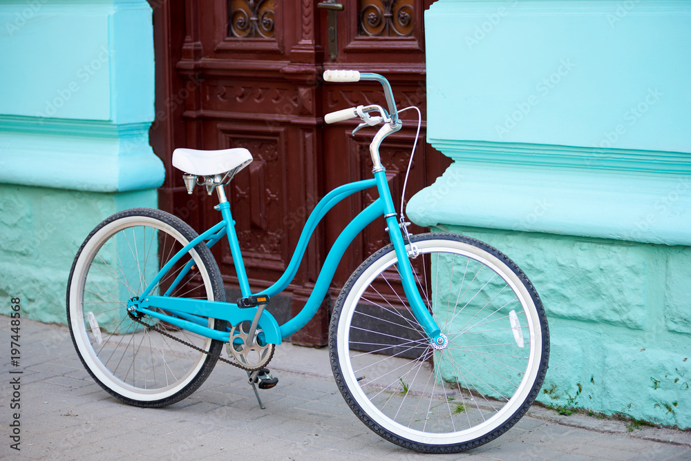 Vintage turquoise bicycle with white elements parked near old building with turquiose walls and brown wooden antique doors.