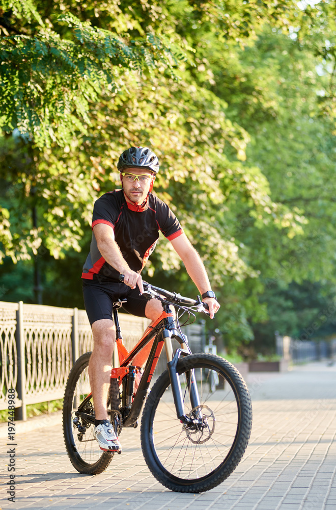 Male biker in cycling sportswear and protective helmet riding on bike near green park. Concept of healthy lifestyle, sport advertising, outdoor activities