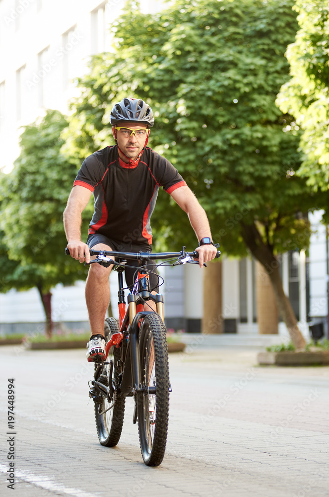 Obraz premium Man bicyclist in cycling garment and protective helmet, looking at camera, riding bike down empty city center alley surrounded by green trees. Sportsman training outdoors improving skills.