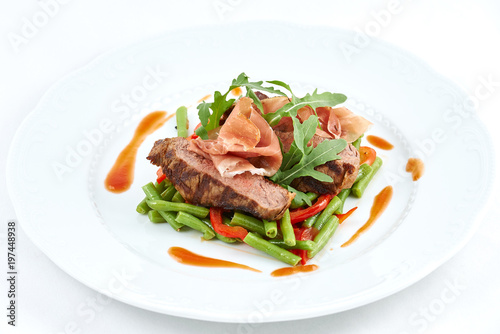 Grilled beef with pork slices and boiled pepper, green beans, fresh arugula and sauce on a white plate.
