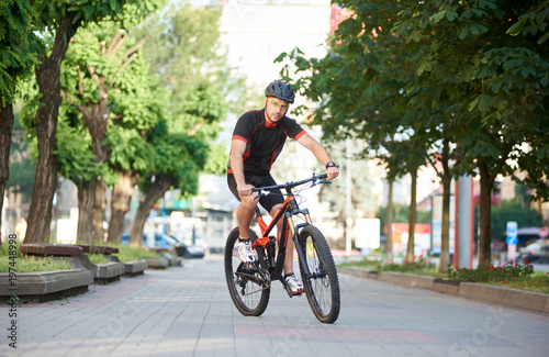 Male cyclist in professional cycling sportswear and protective helmet riding on bike along empty city streets, green trees around. Sportsman performing morning outdoor exercise and training.