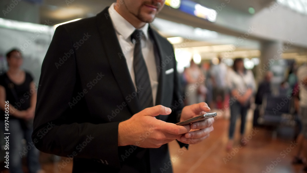 Businessman standing in airport hall, typing a message on phone, business trip