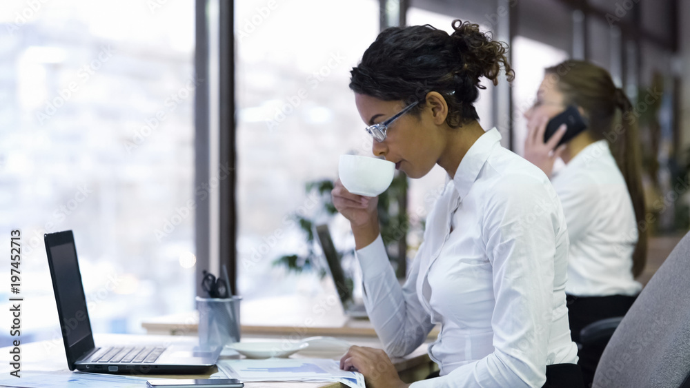 Attractive female office worker drinking cup of coffee and reading charts Attractive female office worker drinking cup of coffee and reading charts