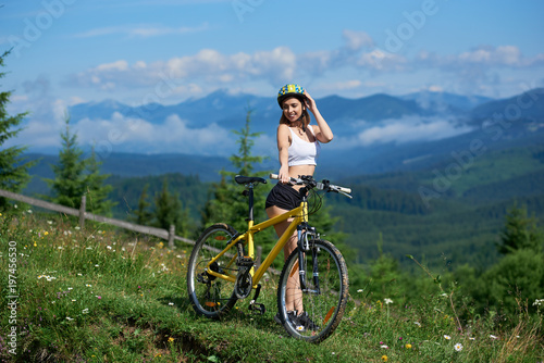 Sporty woman standing on rural trail in mountains with yellow bicycle, wearing helmet, enjoying valley view and foggy mountains on the background in morning. Outdoor lifestyle activity.