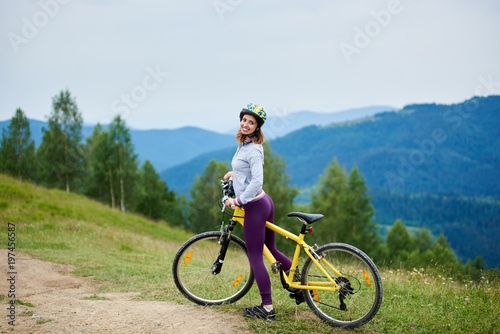 Active smiling female with yellow bicycle on a rural trail on the high mountain range, wearing helmet. Mountains, forests on the background. Outdoor sport activity, lifestyle concept