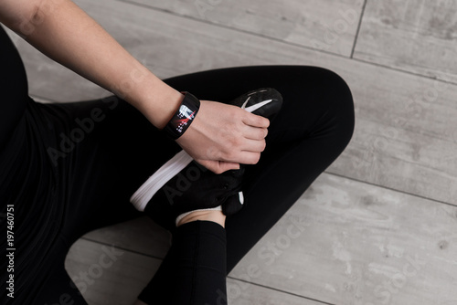 A woman's hand with a smart watch. Holding the foot for foot. Close-up. Black sportswear. The lotus position. Black sneakers with white soles. Wooden floor.