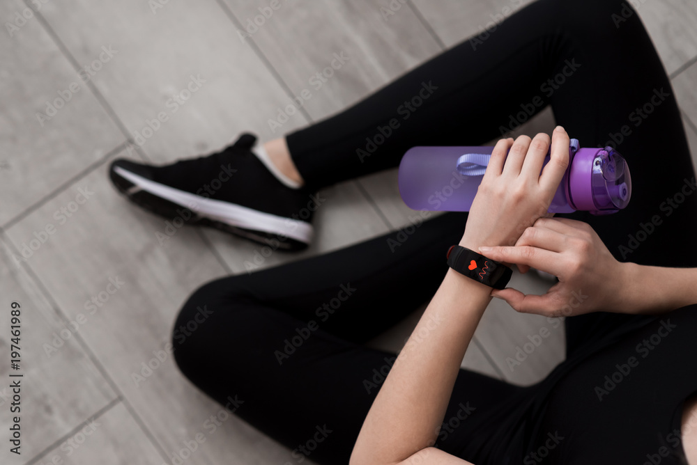A woman's hand with a smart watch. Bottle with water in hand. Heart Rate Measurement. Close-up. Sitting on the floor. Black sportswear.  Black sneakers with white soles. Wooden floor.