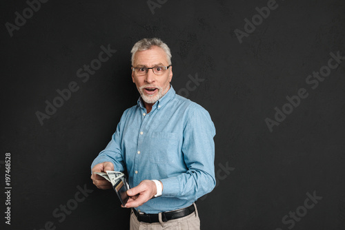 Portrait of adult man 60s with grey hair and beard posing on camera and holding wallet with money, isolated over black background