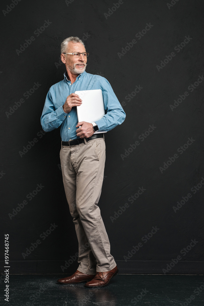 Full length photo of mature stylish man 60s with grey hair and beard standing and holding silver laptop in hands, isolated over black background