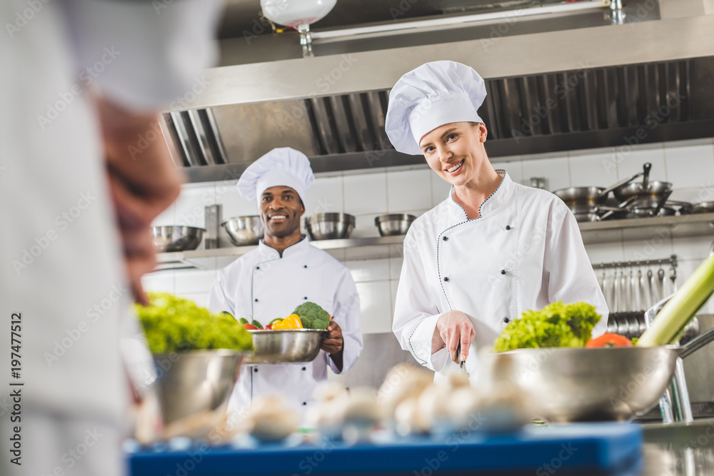 smiling multicultural chefs preparing food at restaurant kitchen and looking at camera smiling multicultural chefs preparing food at restaurant kitchen and looking at camera