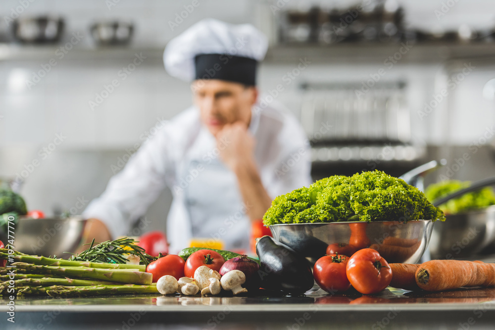 pensive chef at restaurant kitchen with vegetables on foreground pensive chef at restaurant kitchen with vegetables on foreground