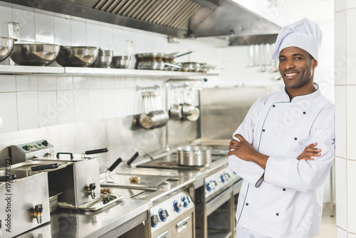 smiling african american chef standing with crossed arms and looking at camera at restaurant kitchen