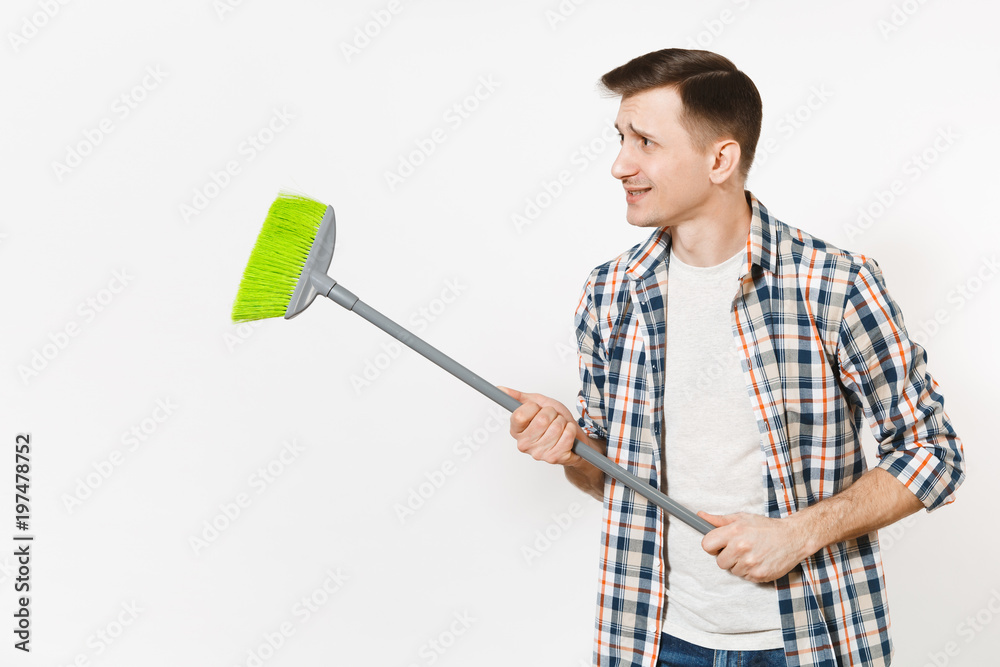 Young strange fun housekeeper man in checkered shirt holding and sweeping with green broom isolated on white background. Male doing house chores. Copy space for advertisement. Cleanliness concept. Young strange fun housekeeper man in checkered shirt holding and sweeping with green broom isolated on white background. Male doing house chores. Copy space for advertisement. Cleanliness concept.