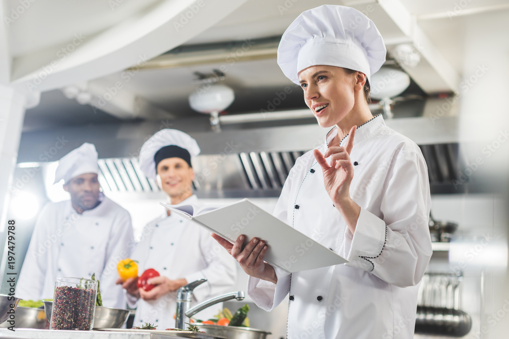 attractive chef holding recipe book and showing idea gesture at restaurant kitchen