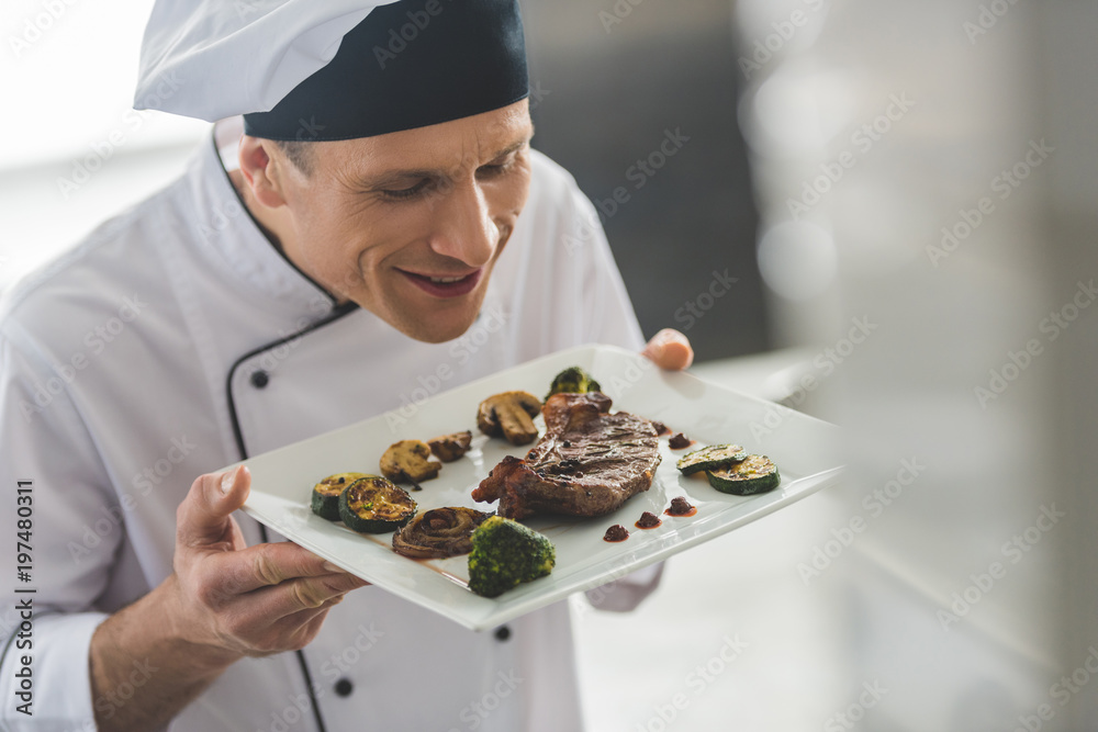 handsome chef sniffing cooked steak with vegetables at restaurant kitchen