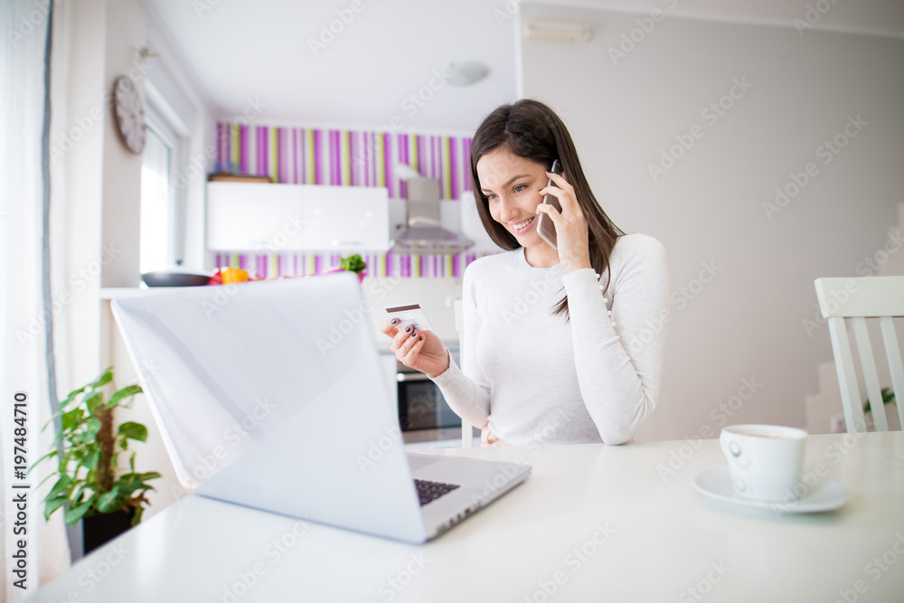 Young beautiful happy girl is checking her credit card info and looking at laptop while drinking coffee and talking on the phone at the kitchen table. Young beautiful happy girl is checking her credit card info and looking at laptop while drinking coffee and talking on the phone at the kitchen table.