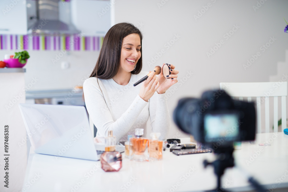 Young beautiful cute girl is sitting at the kitchen table with a laptop on it and showing perfumes and cosmetics to the camera while laughing.