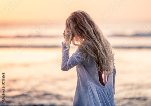 Obraz na plátne Beautiful blonde girl with long hair in short white dress walking at sunset on t