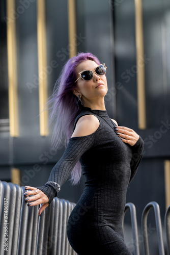 Caucasian white female model standing on the balcony of the building and enjoys weather. Beautiful woman with long purple hair, in a gray dress resting in the city, modern fashion concept.