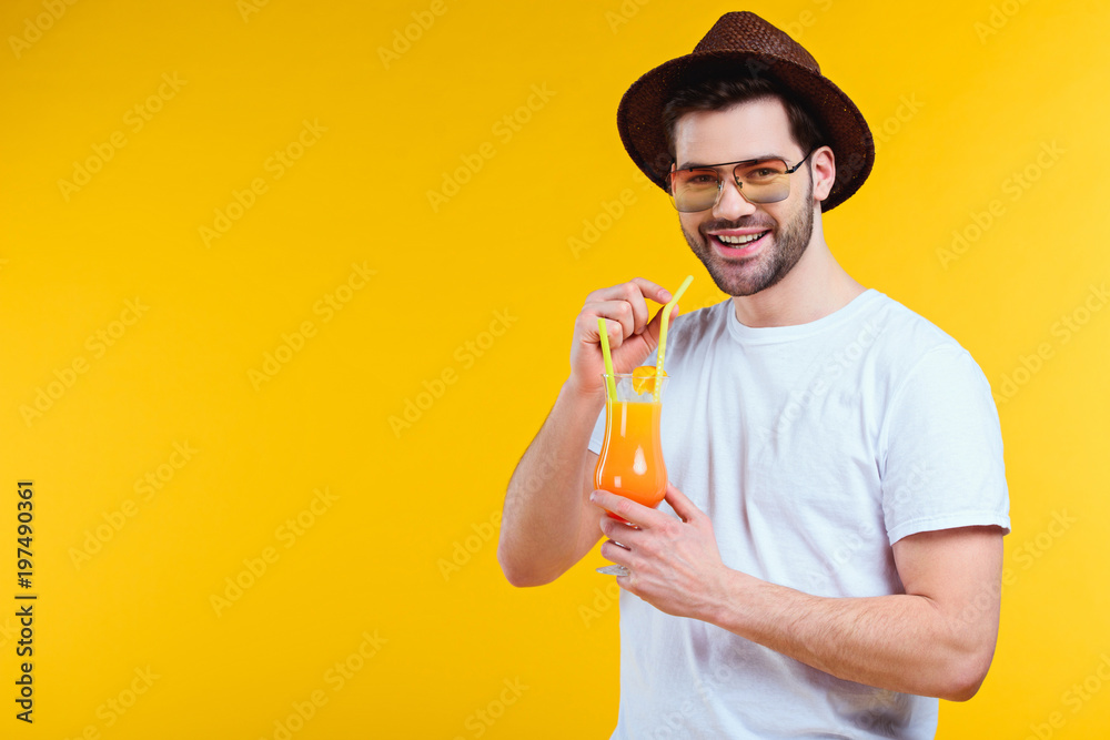 handsome young man in hat and sunglasses drinking summer cocktail and smiling at camera isolated on yellow handsome young man in hat and sunglasses drinking summer cocktail and smiling at camera isolated on yellow