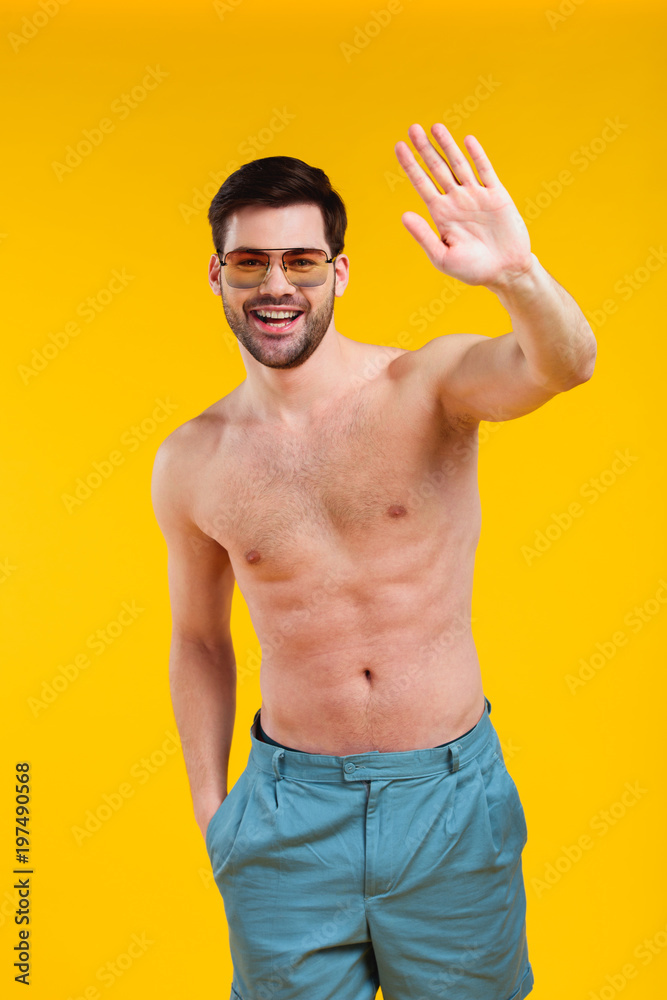 handsome shirtless young man in shorts and sunglasses waving hand and smiling at camera isolated on yellow