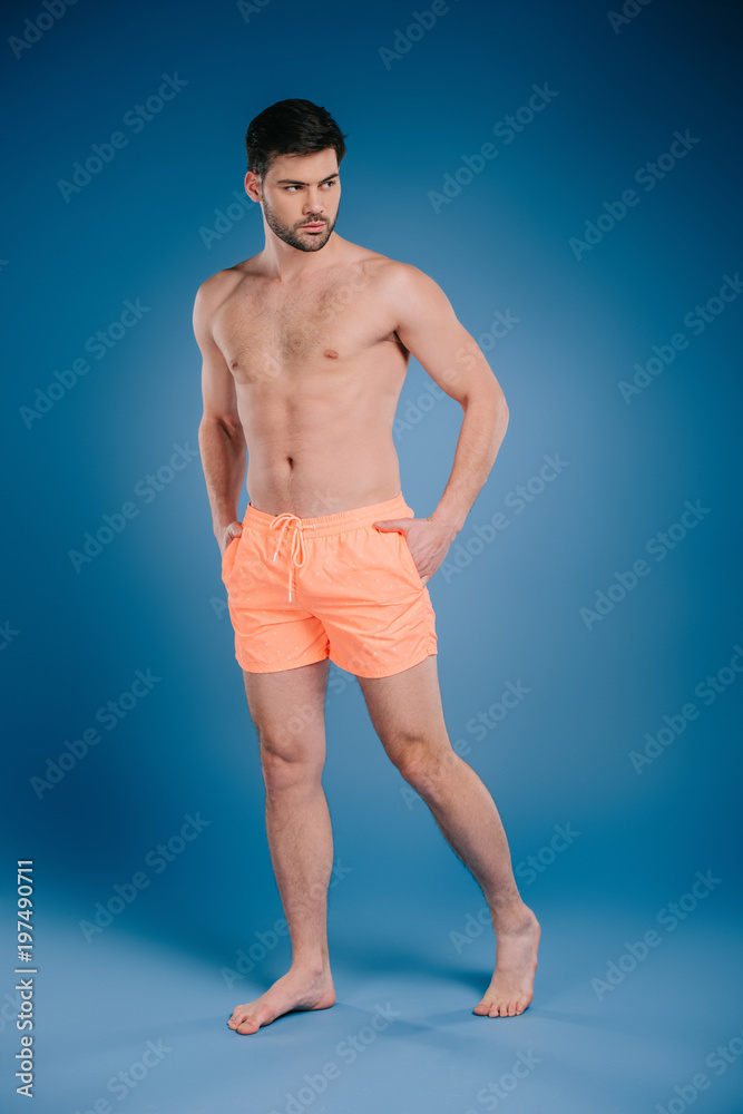 full length view of handsome barefoot man in shorts standing with hands in pockets and looking away on blue