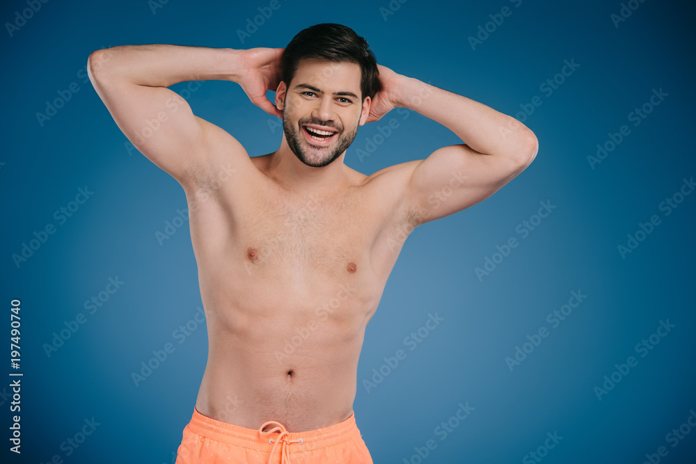 handsome shirtless young man standing with hands behind head and smiling at camera on blue