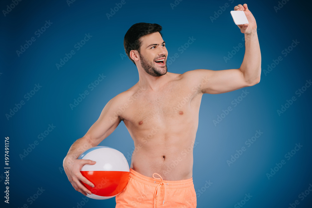 smiling young man in shorts holding beach ball and taking selfie with smartphone on blue