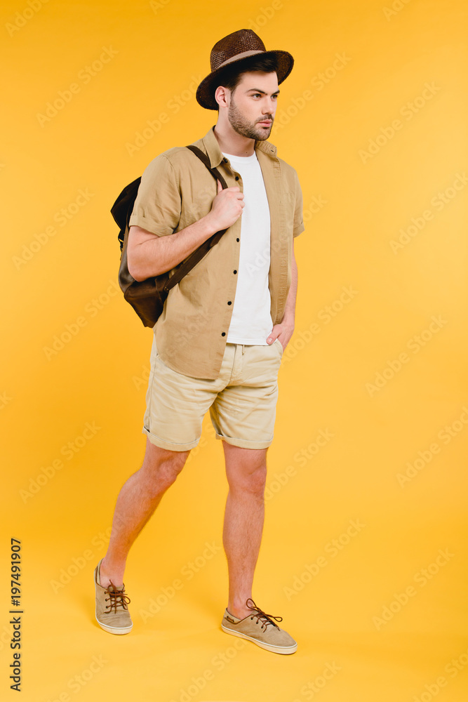 full length view of young man in shorts and hat holding backpack and walking isolated on yellow full length view of young man in shorts and hat holding backpack and walking isolated on yellow