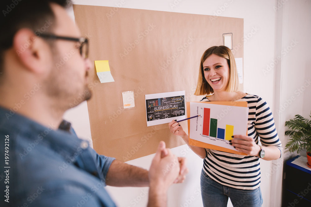 Two young business people consulting about a analyse in front of the poster with a business plan. Young smiling woman showing company progress to man. Two young business people consulting about a analyse in front of the poster with a business plan. Young smiling woman showing company progress to man.