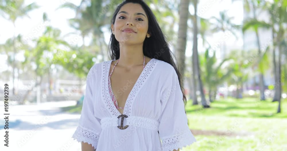 Amazing ethnic woman wearing bikini and white dress smiling at camera standing on background of green sunny park. Amazing ethnic woman wearing bikini and white dress smiling at camera standing on background of green sunny park.