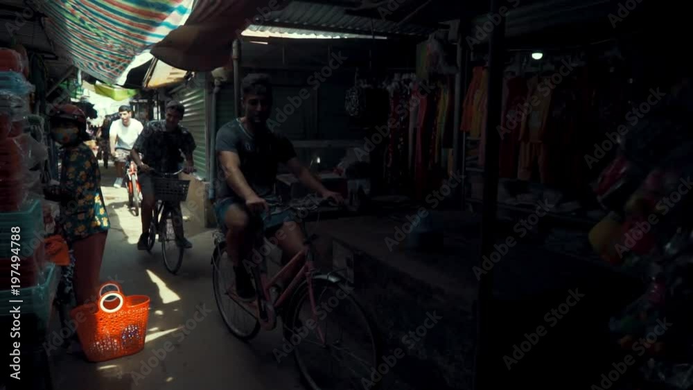 A group of tourist friends ride bicycles through a dark and narrow marketplace in Ho Chi Minh City A group of tourist friends ride bicycles through a dark and narrow marketplace in Ho Chi Minh City