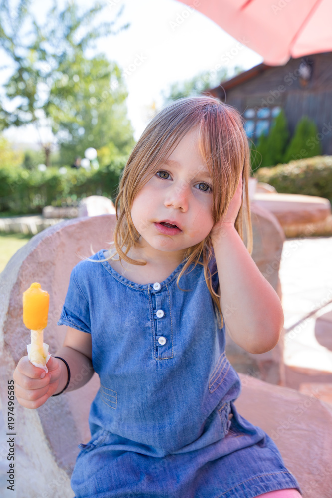 portrait of four years old blonde girl looking with blue denim dress, holding colorful orange or yellow ice lolly or popsicle, sitting in public park
