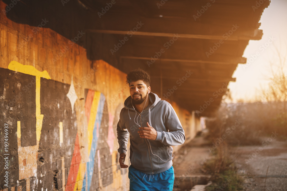Portrait of active motivated afro-american young attractive athletic man with earphones running inside of the abandoned place. - obrazy, fototapety, plakaty 