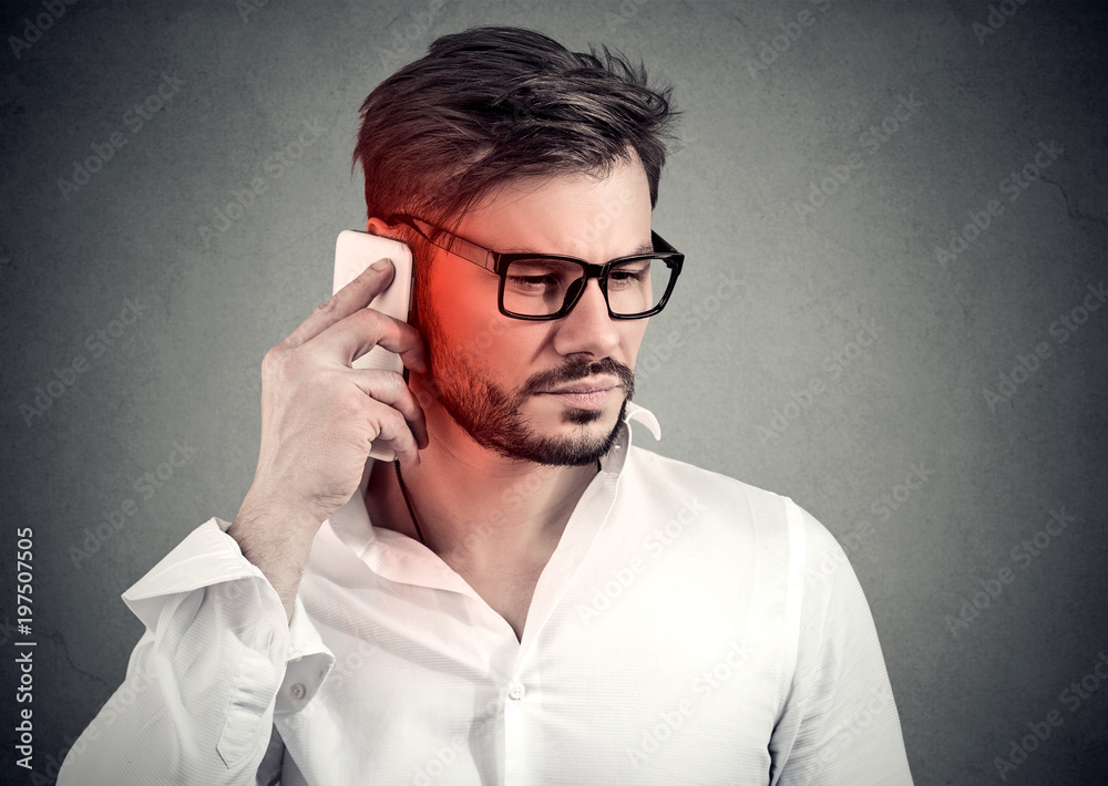 Obraz premium Man on the phone with headache. Upset unhappy guy talking on a cellphone on gray background. Negative emotion face expression