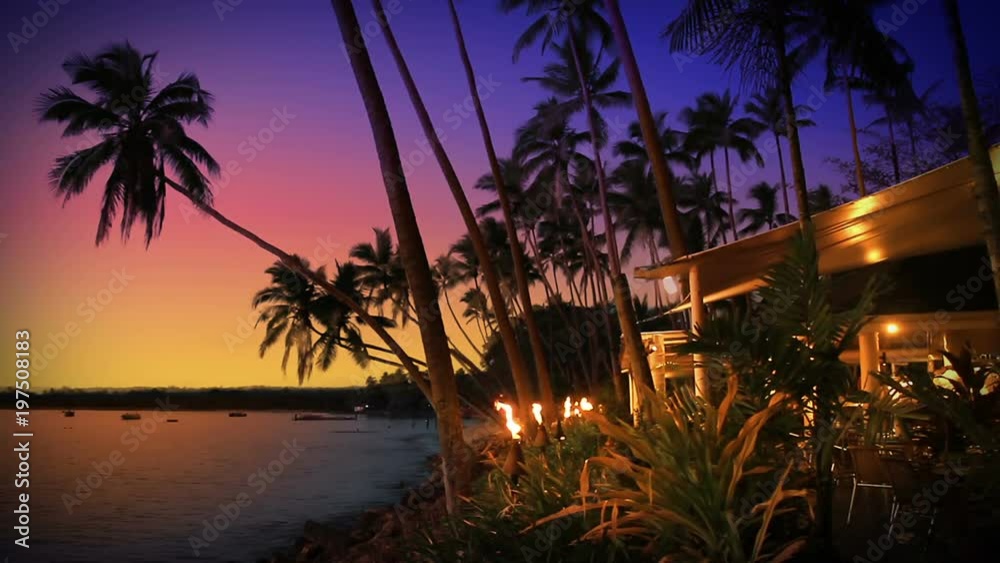Beach-side restaurant / cafe / bar lit by flaming torches, at sunset in tropical Fiji. Idyllic romantic summer night dining under a purple sky.