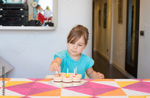 three years old blonde child putting candles on birthday cake on colorful tablecloth at home 