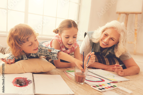 Feeling comfortable. Joyful nice delighted kids holding brushes and painting while lying on the floor