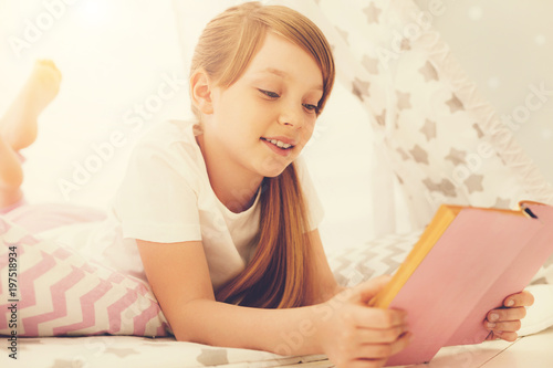 Time for adventures. Happy positive cheerful girl lying on the bed and reading a story while having a pleasurable time