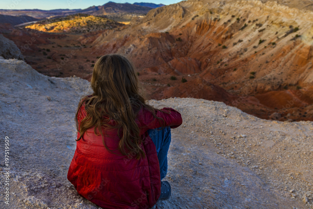 A child in a red jacket sitting and enjoying the view of a desert canyon in the evening