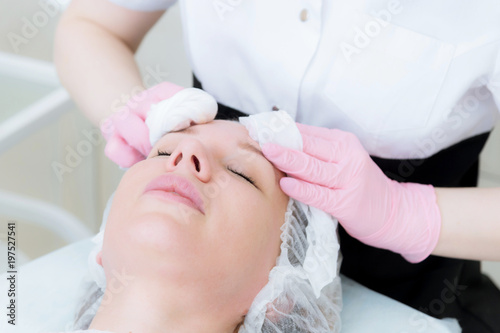 A close-up of the cleaning procedure in the office of cosmetology. The hands of the cosmetologist in pink gloves are removed from the face of a young girl with a sponge cleansing the mask