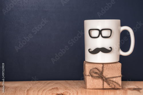 cup with mustache, glasses, gift box over black chalkboard, holiday concept of Fathers Day