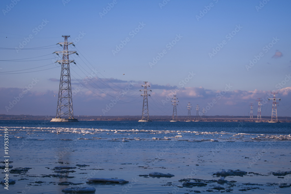 the electric transmission line passing through the river against the sky and the visible opposite shore
