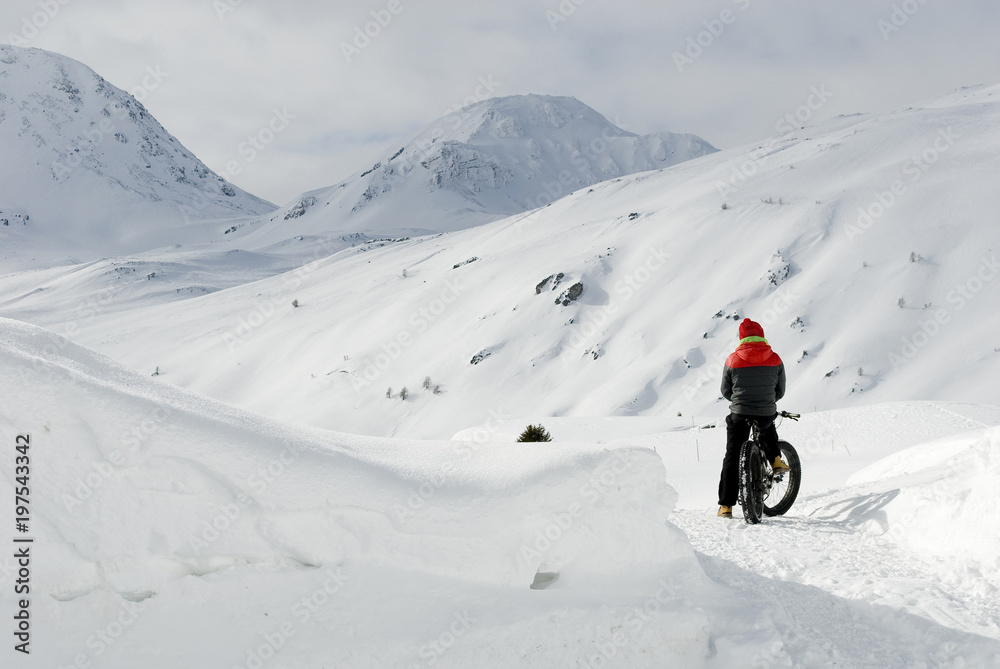 man use electric bicycle, e-bike, ebike, snow covered road, look mountains and horizon, bike with wide wheels to go on snow, called fatbike, winter, cold, alps, freeride, Simplon Pass, Switzerland